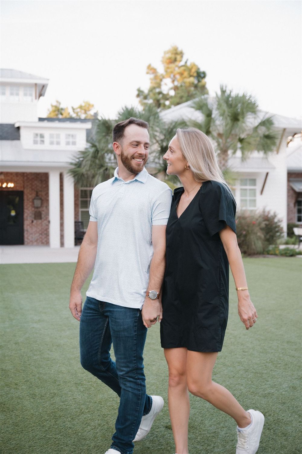 A couple walks hand in hand on a green lawn in front of a white house, sharing a sweet moment together.