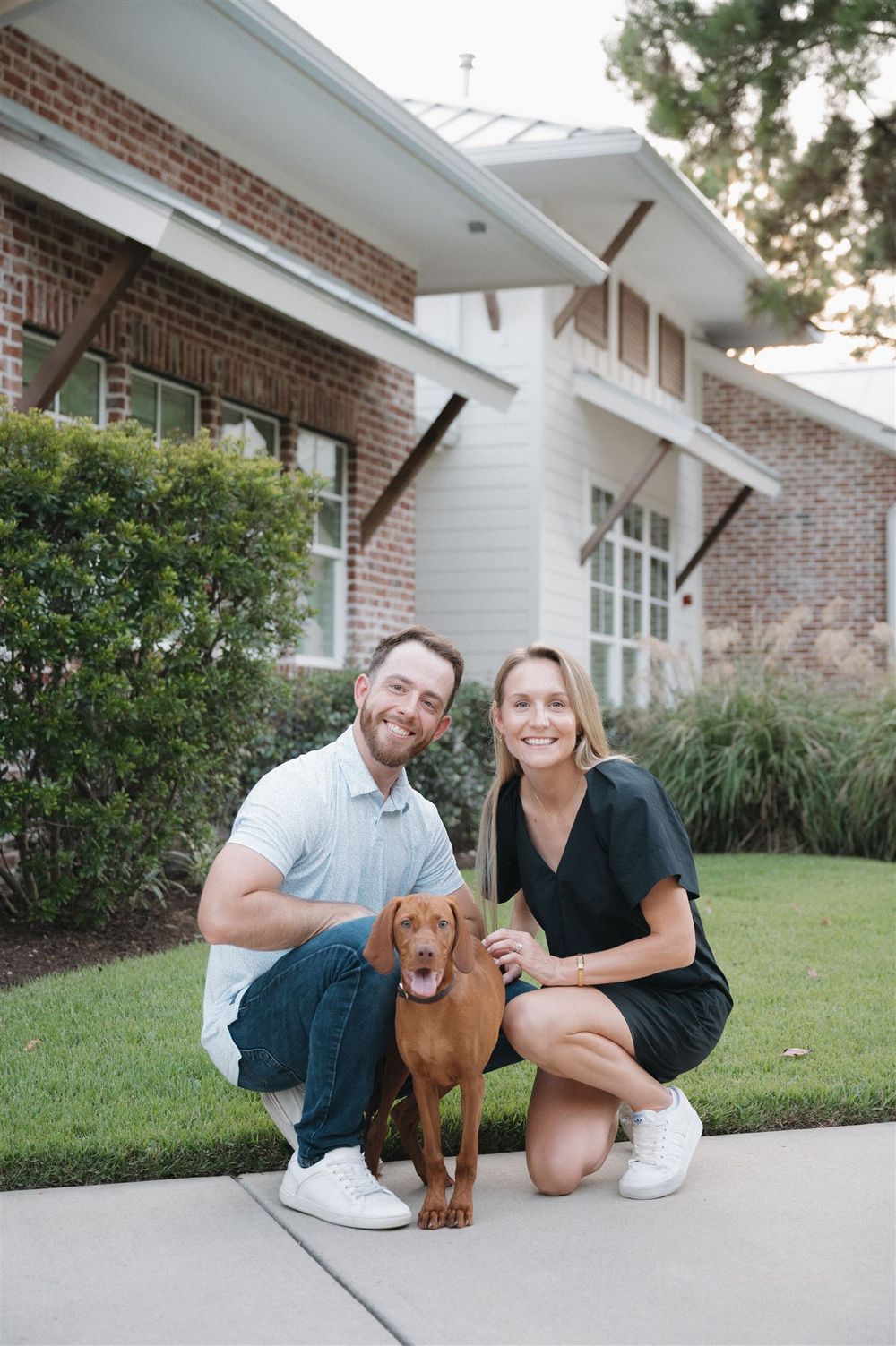 A red Vizsla dog sits with two people on a pathway outside a brick building with white trim.
