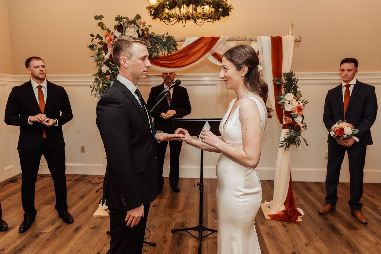 A wedding ceremony in a classic reception room with decorative floral arrangements and elegantly dressed attendants.