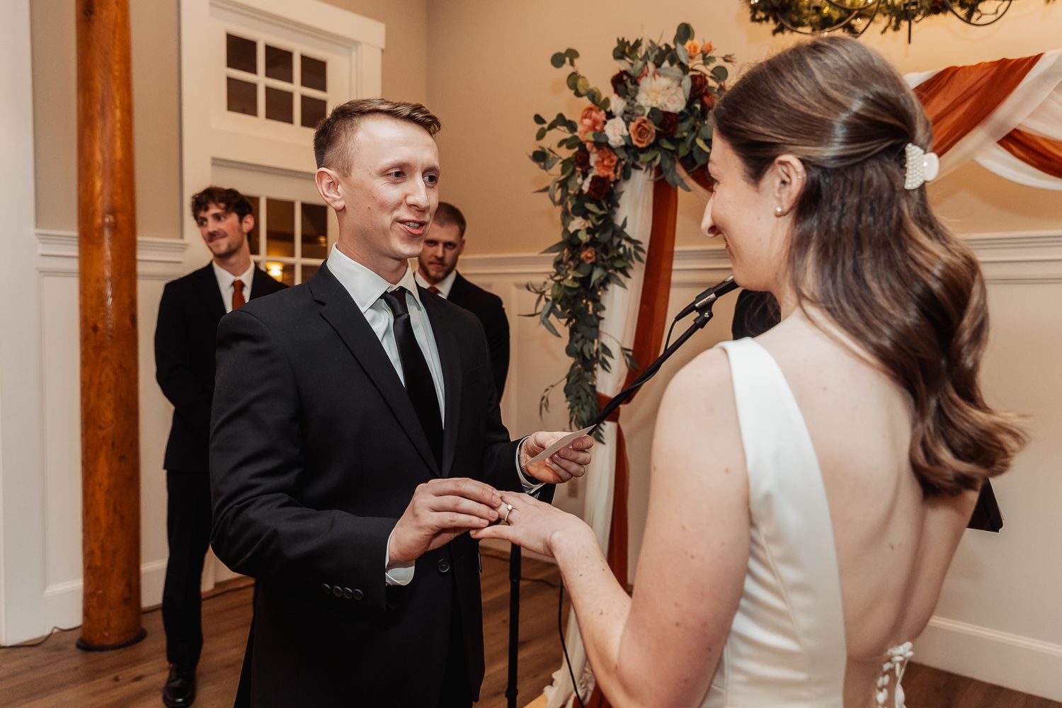 Wedding ceremony exchange of rings in an intimate indoor setting with decorative floral arch.