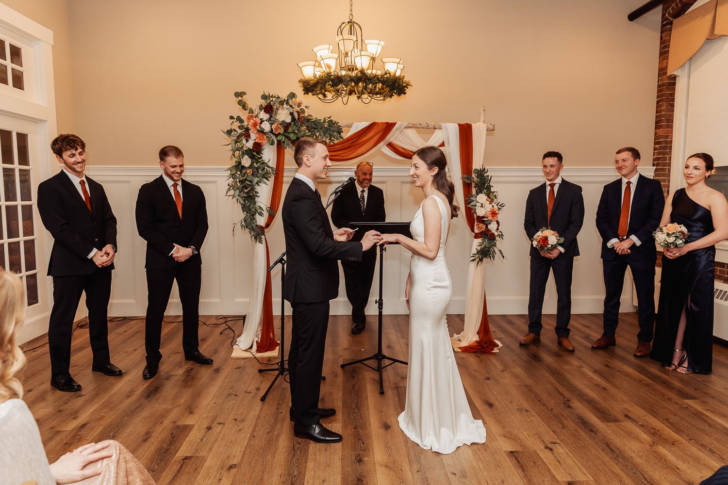 Wedding ceremony taking place in an elegant indoor venue with copper arch, greenery and wooden floors.