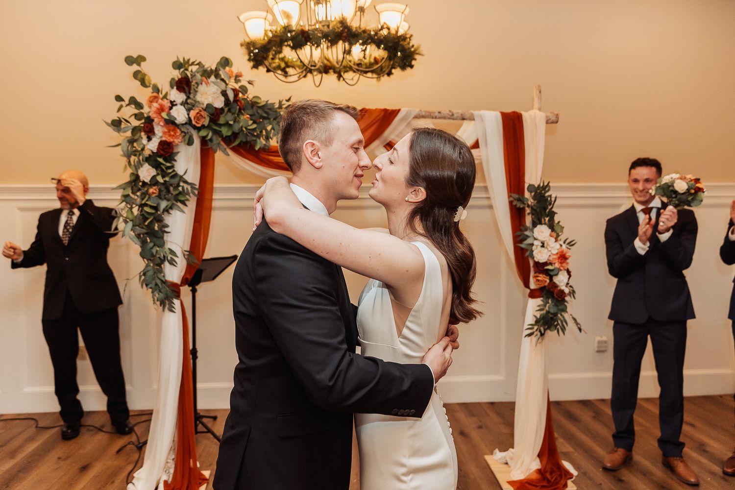 Two people share a first dance as newlyweds under ornate chandelier with floral arch and onlookers at indoor venue.
