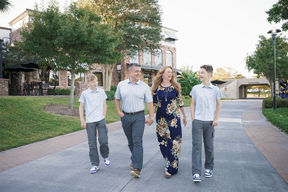 A group of four people stroll together along a paved walkway on a sunny day with landscaped grounds and buildings behind them.