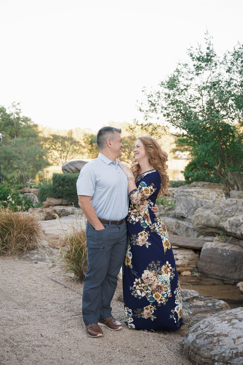 A romantic couple embraces on a rocky path during sunset, with the woman wearing a navy floral maxi dress.