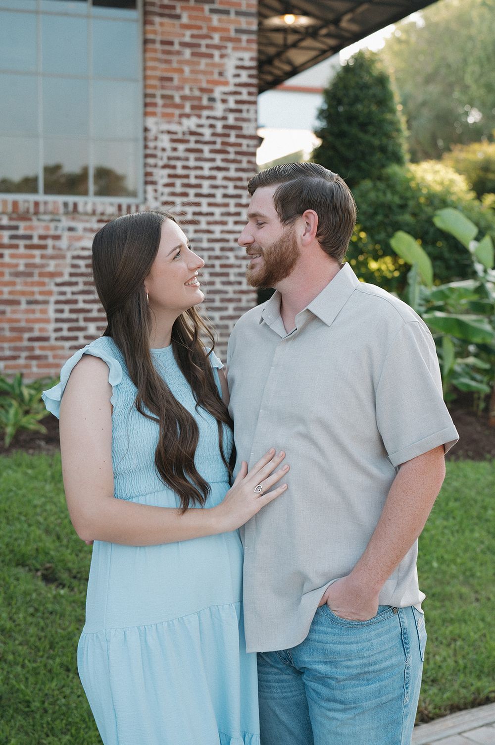 A couple shares a tender moment together outside a brick building, with her wearing a light blue dress and him in grey.