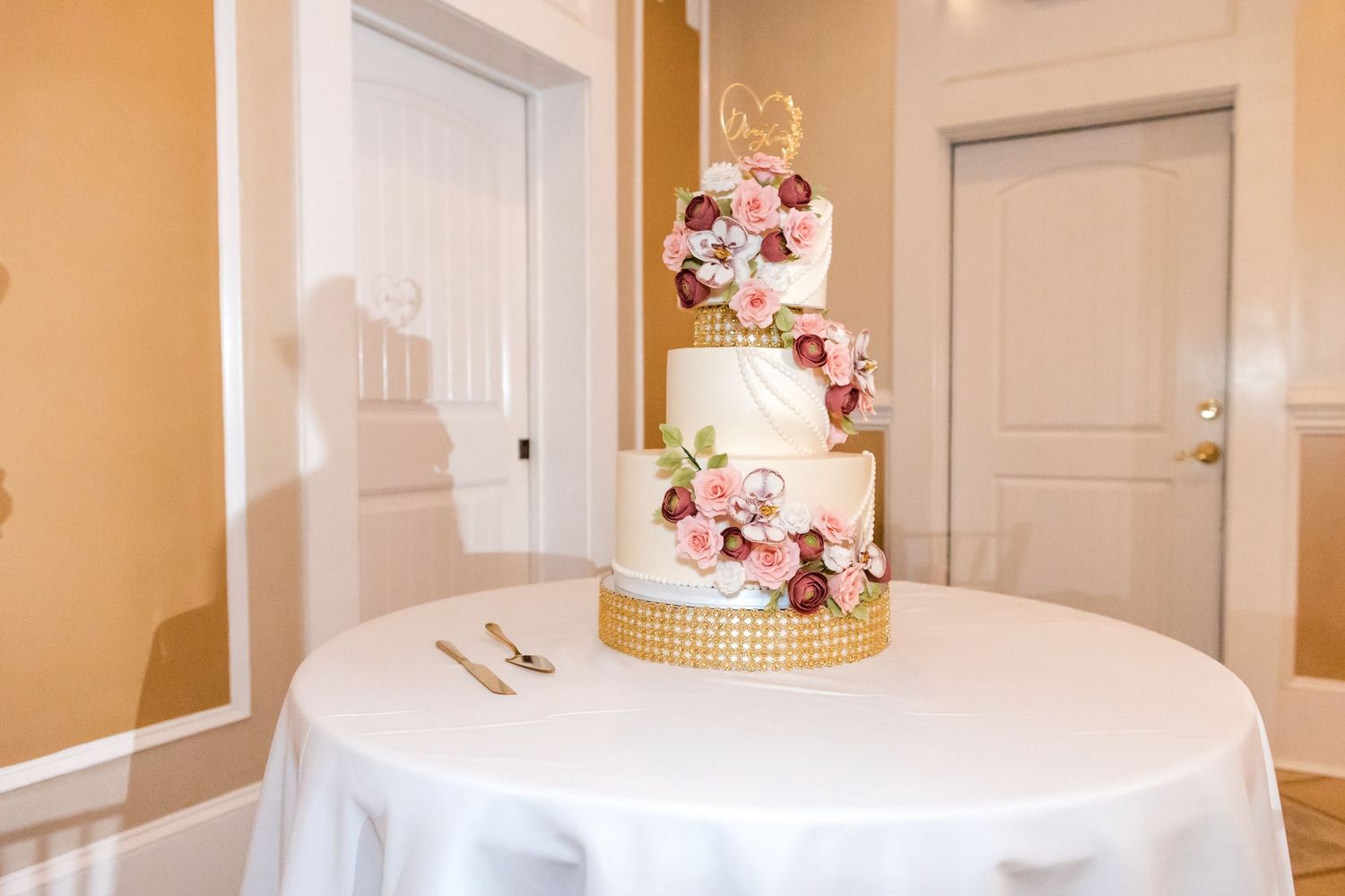 Elegant three-tier white wedding cake decorated with pink and burgundy roses on a white tablecloth.
