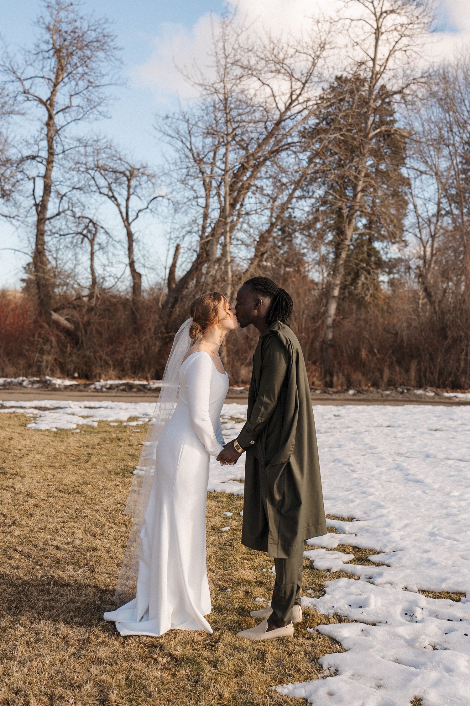 A couple stands together in a snowy winter landscape with bare trees, one wearing a white dress and one in dark coat.