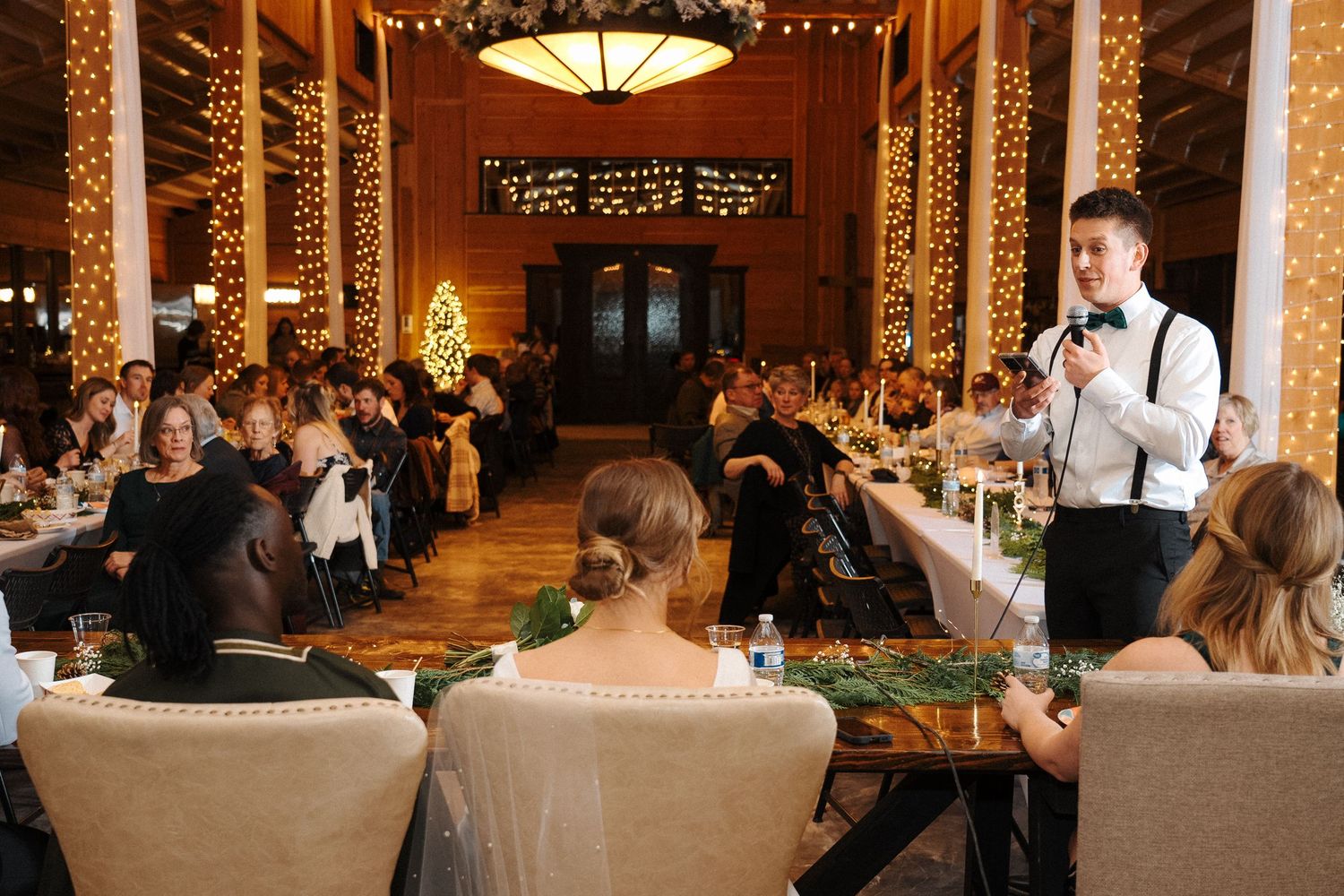 Server addresses wedding guests seated in an elegant rustic venue with wooden beams and ambient lighting during reception.
