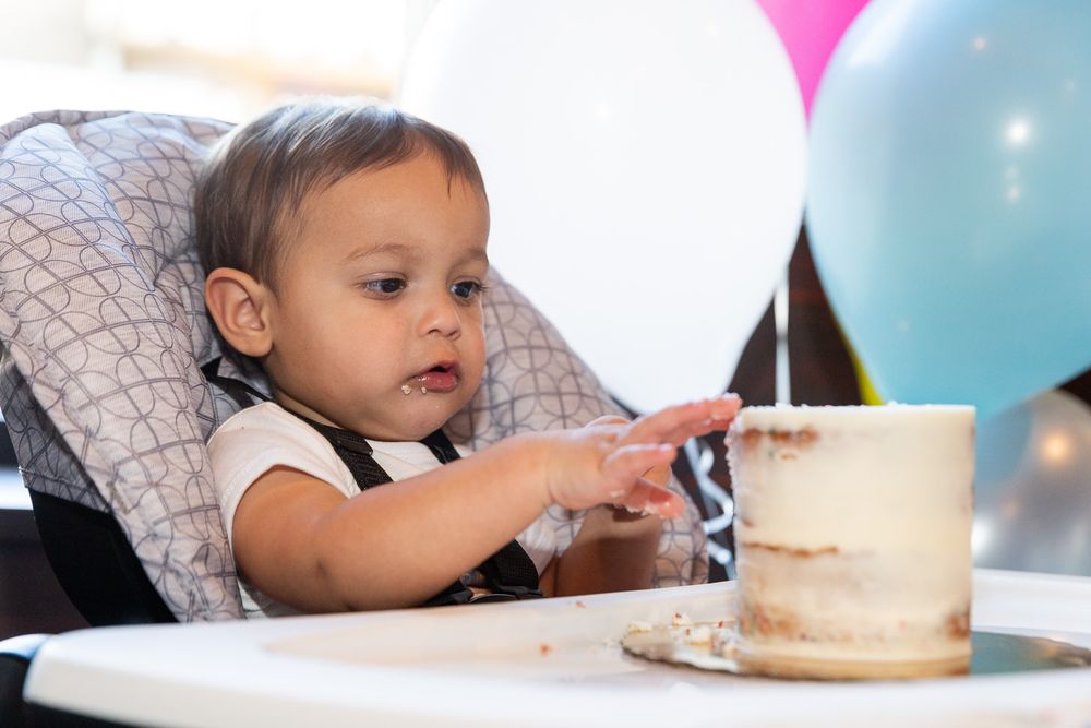 Baby in a highchair reaching for a small white cake with balloons in the background.