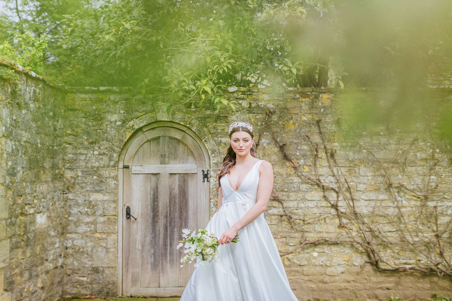 A bride in a wedding dress poses in front of a Cotswolds stone wall with an arched wooden door. Cornwell Manor Hayley Elizabeth Photography