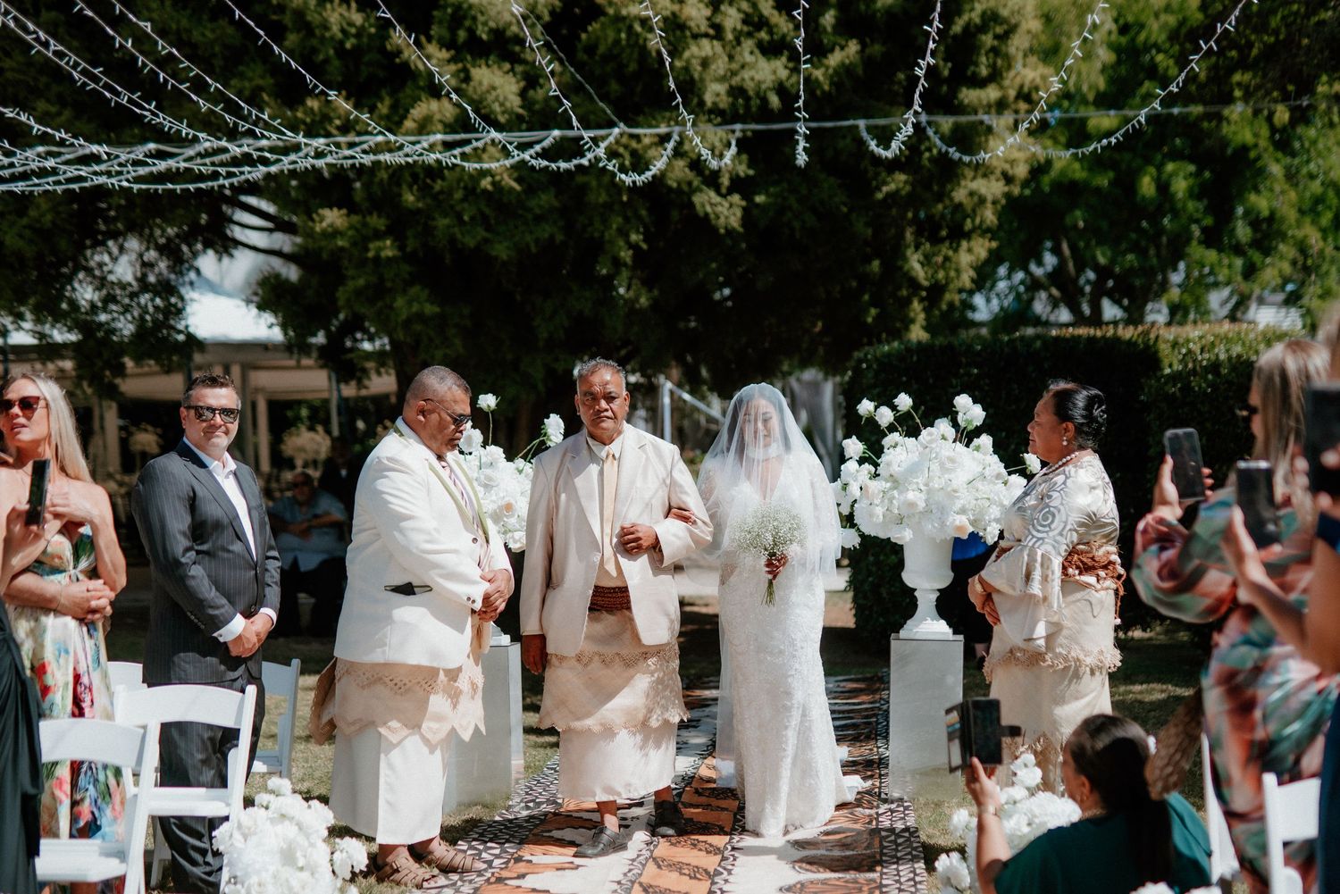 Outdoor wedding ceremony under string lights with guests in cream-colored attire.