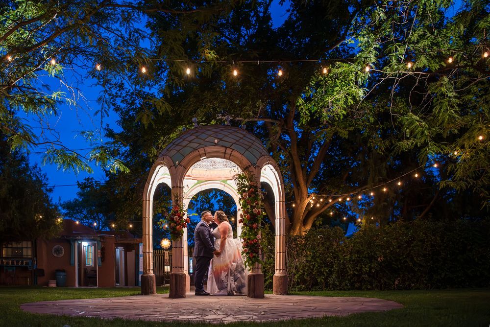 A couple shares a romantic moment under an illuminated stone archway at dusk with string lights overhead.