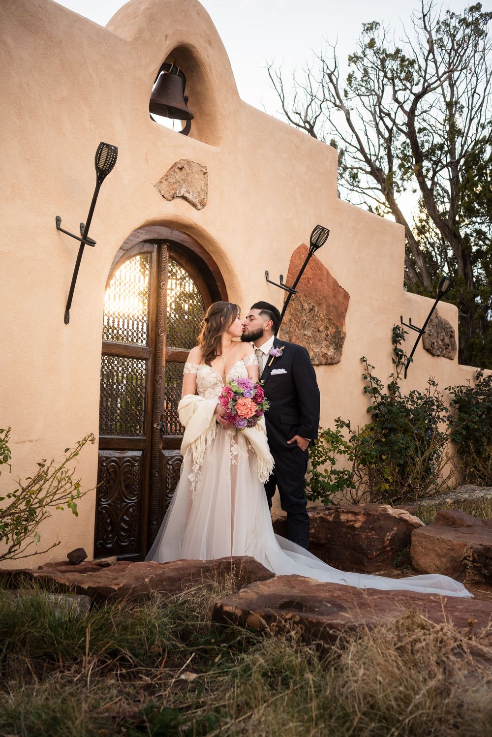 A bride and groom embrace in front of a stunning adobe-style entrance with rustic metal lanterns and an ornate wooden door.
