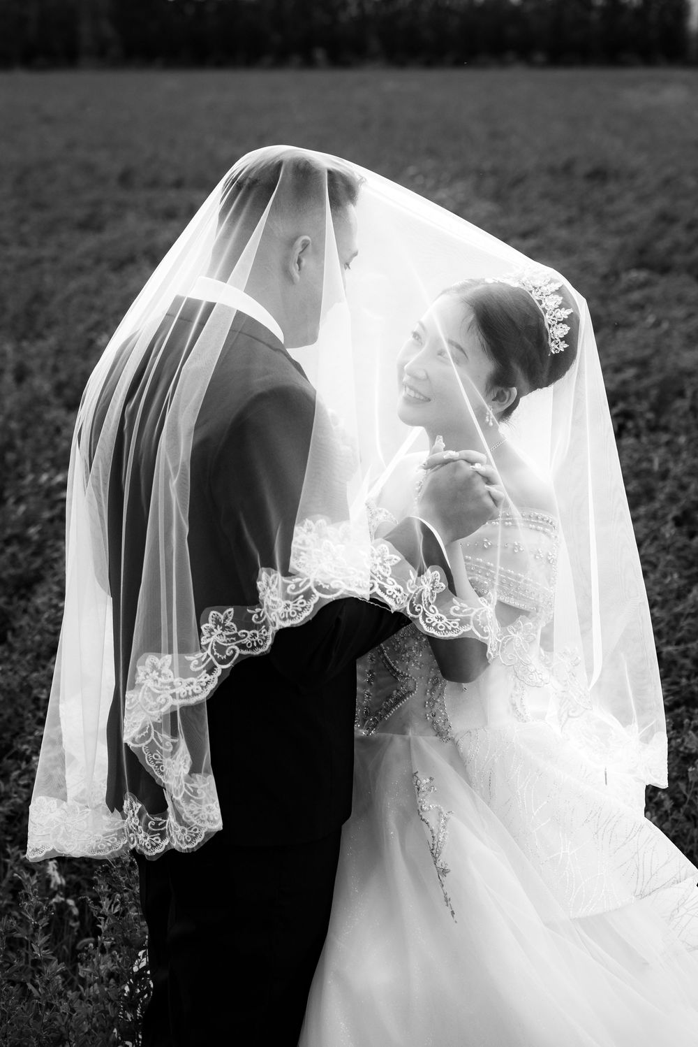 A romantic black and white wedding photo showing a couple under an elegant lace veil in an outdoor setting.