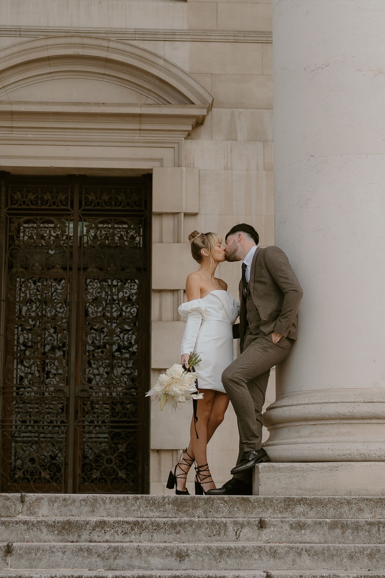 Couple shares a romantic moment on stone steps in front of ornate wooden doors, bride in short dress and groom in gray suit.
