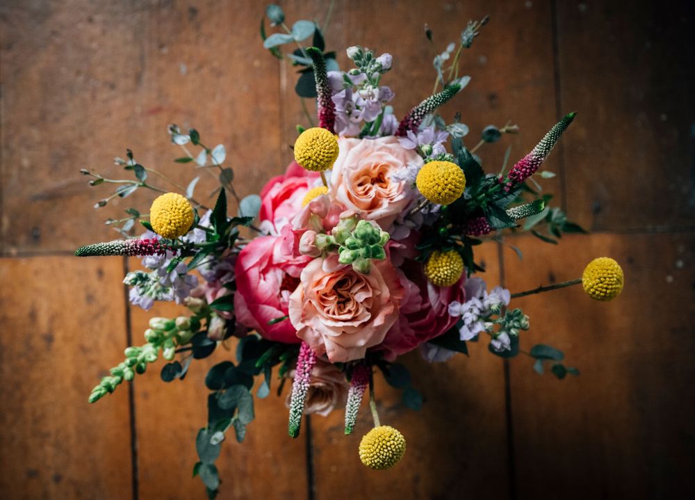 A rustic floral arrangement with pink peonies, yellow craspedia, and purple wildflowers against a weathered wooden background.