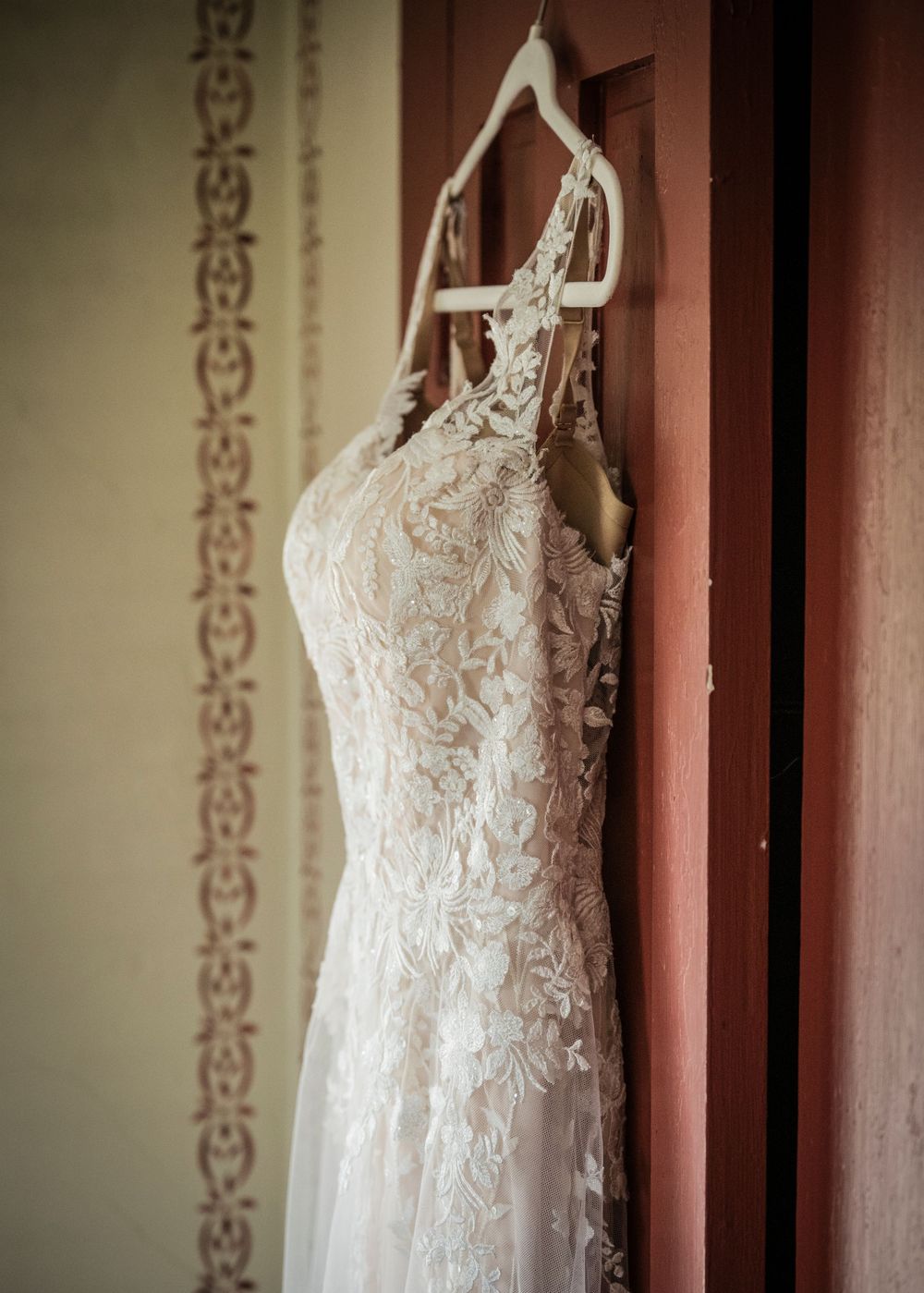 Elegant lace wedding dress with intricate floral details hanging on display against a terracotta-colored wall.