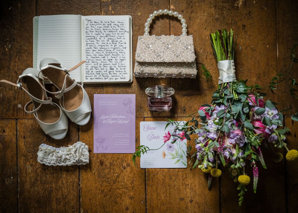 Wedding accessories including lace shoes, vintage handbag, written cards and a colorful floral bouquet arranged on wooden floor.