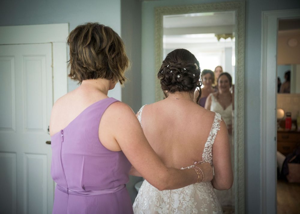 A touching behind-the-scenes moment of wedding preparation as a helper assists with a dress in a lavender gown.
