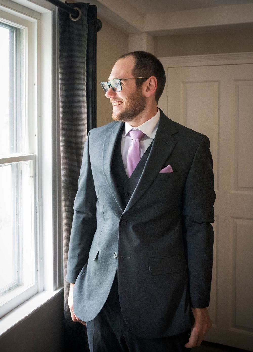 A person in a gray suit with pink tie and pocket square gazes out a bright window in a formal indoor setting.