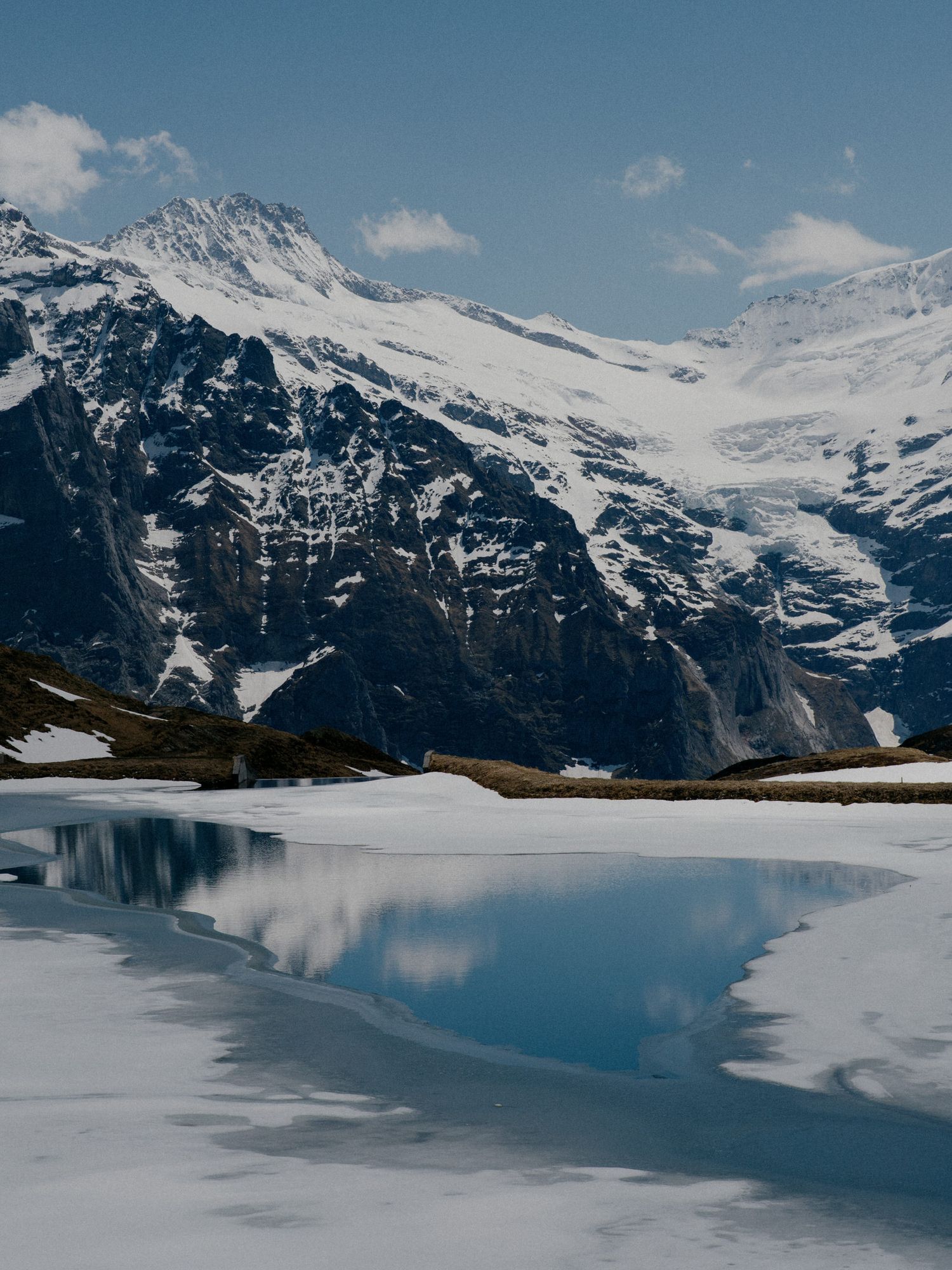 Snow-capped mountain peaks reflect in a partially frozen alpine lake under blue skies in a stunning winter landscape.