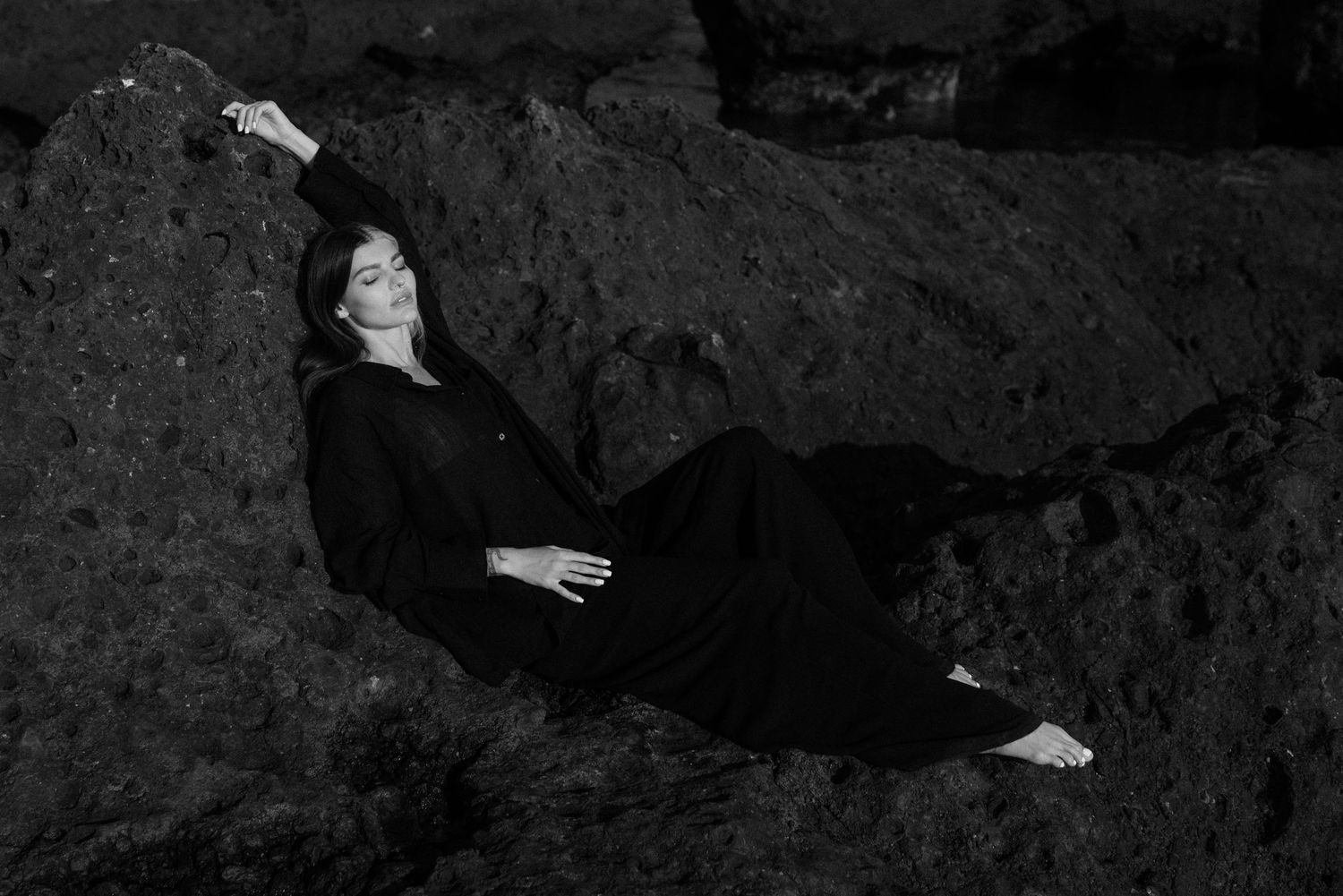 A person in a black outfit reclines dramatically against dark rocks in a black and white artistic photograph.