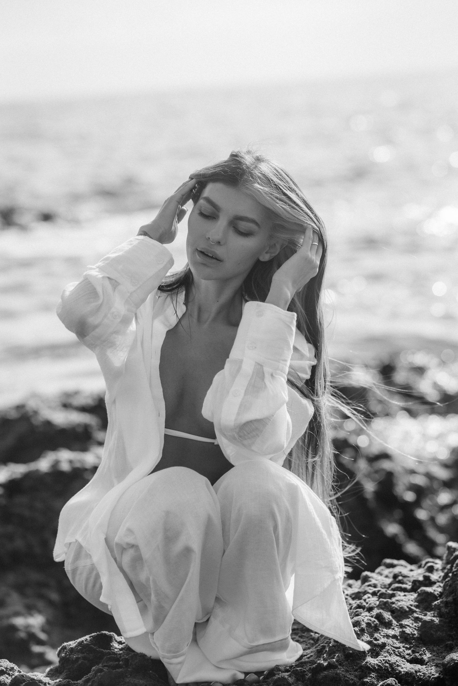 Black and white image of someone in a white flowing shirt and pants crouching on coastal rocks at the beach.