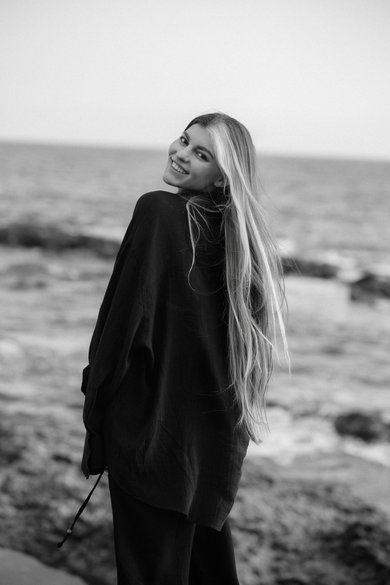 Black and white photo series of a person with long blonde hair standing by a rocky ocean shore in a black sweater.