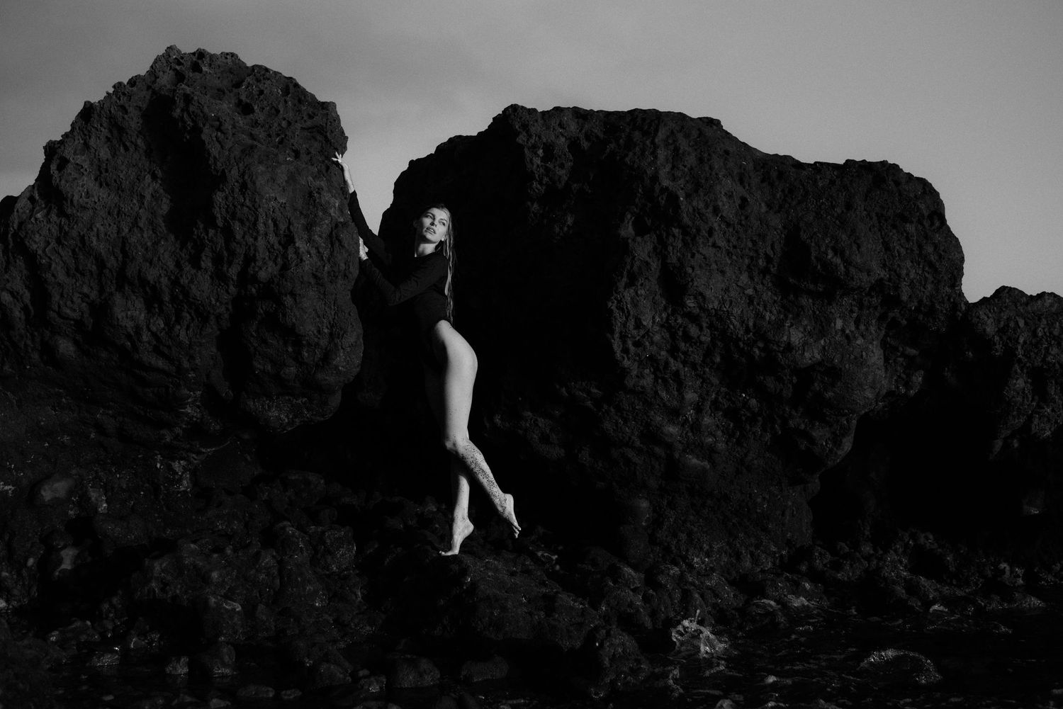 A black and white artistic photo shows a figure posing dramatically against dark coastal rock formations.
