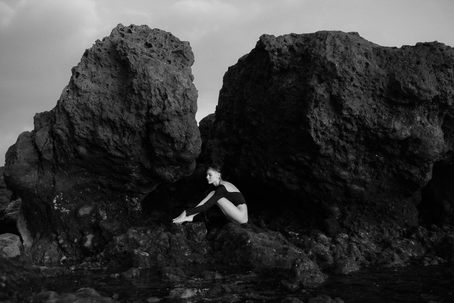 A black and white photograph shows a figure seated between dramatic coastal rock formations at the water's edge.