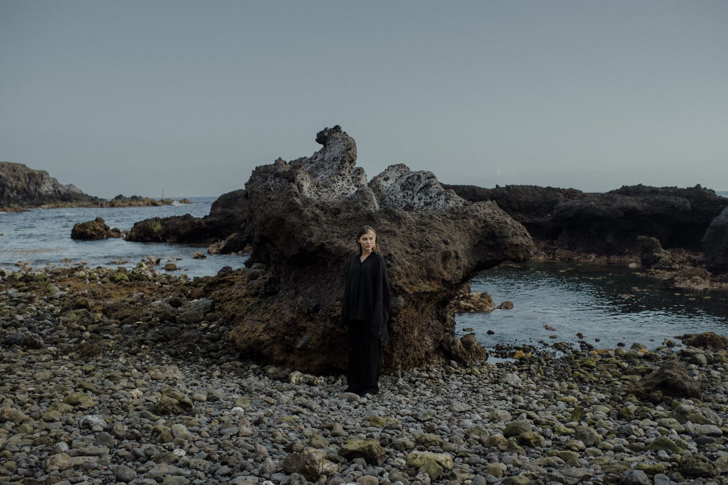 A person in dark clothing stands on a rocky coastline with dramatic rock formations and ocean waves in moody lighting.