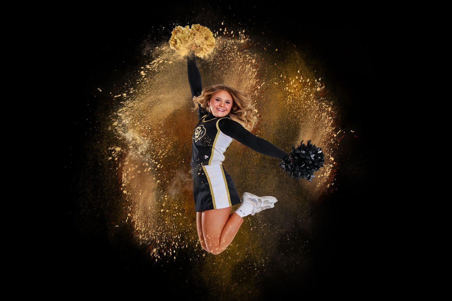 Cheerleader in dark uniform leaps through dramatic gold powder explosion effect against black background.