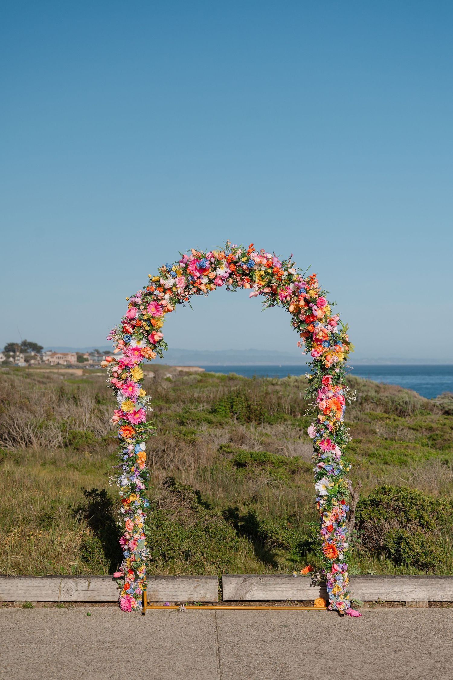A floral wedding arch decorated with pink and colorful flowers stands against a coastal landscape with blue sky and ocean view.