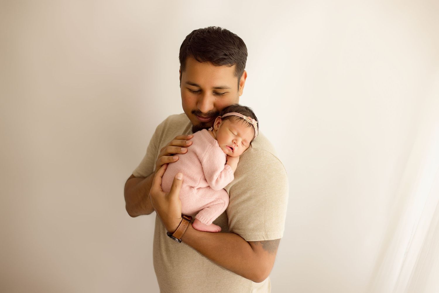 A tender moment between a parent wearing a beige shirt holding a newborn baby in pink against a light background.