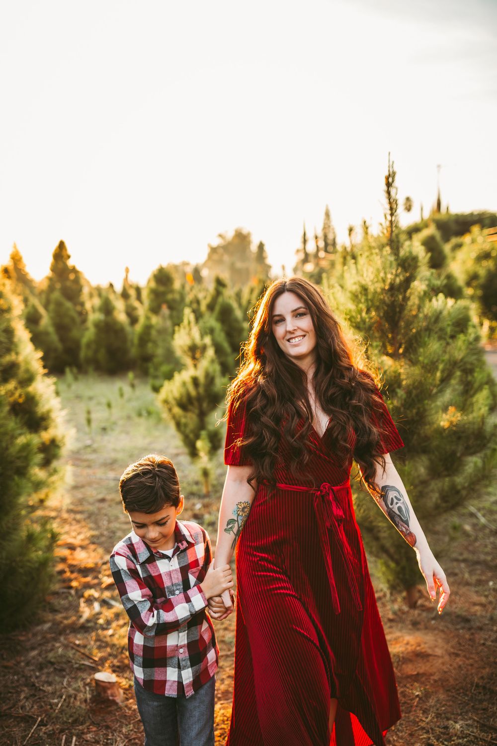Mother and child sharing a playful moment among evergreen trees at sunset in a Christmas tree farm.