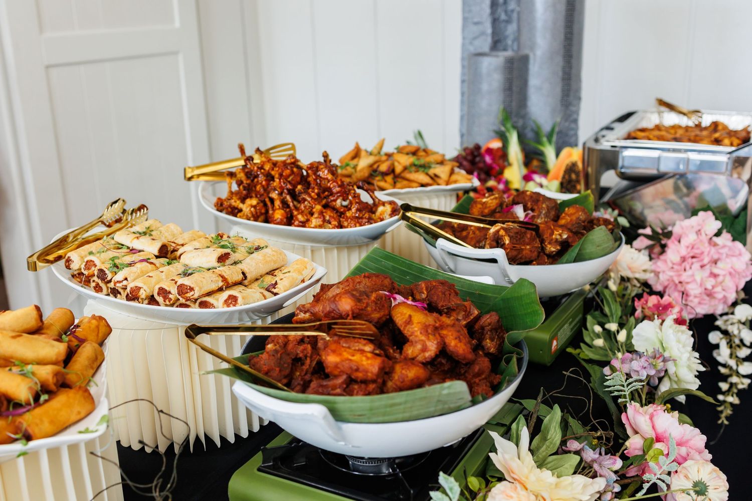 Buffet table spread with fried appetizers, chicken wings and pink floral decorations arranged on white tablecloth.