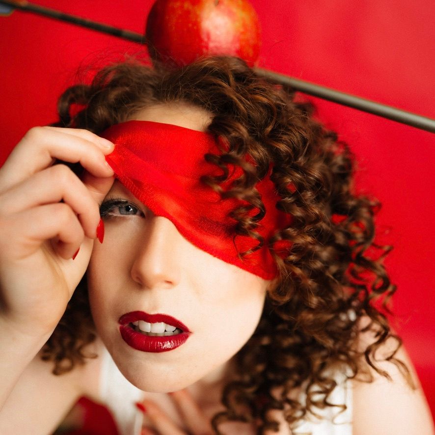 Glamorous portrait with red blindfold, curly brown hair, and bright red lipstick against vibrant red background.