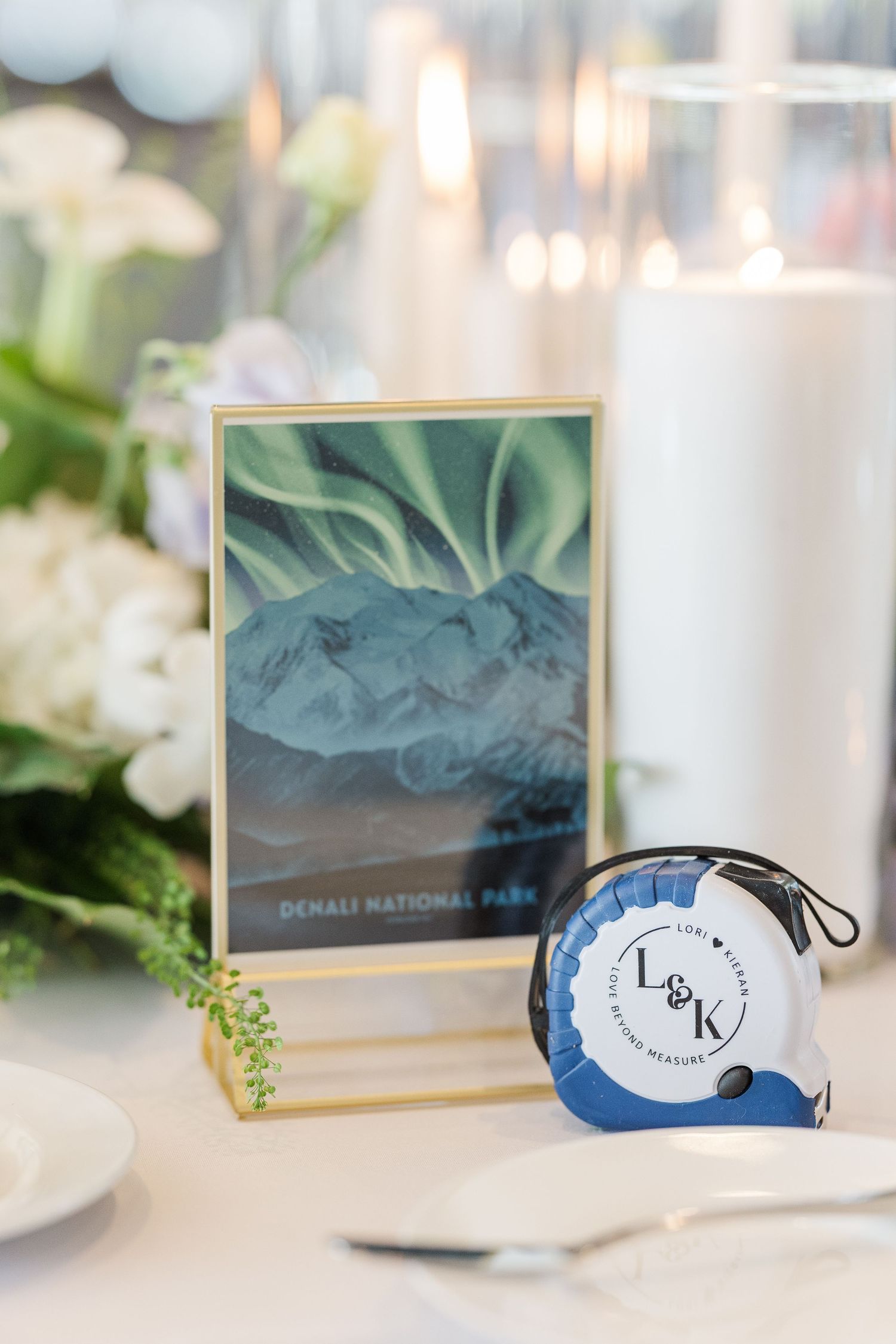 Wedding table decor features a framed Denali National Park print and blue monogrammed favor against white floral arrangements.