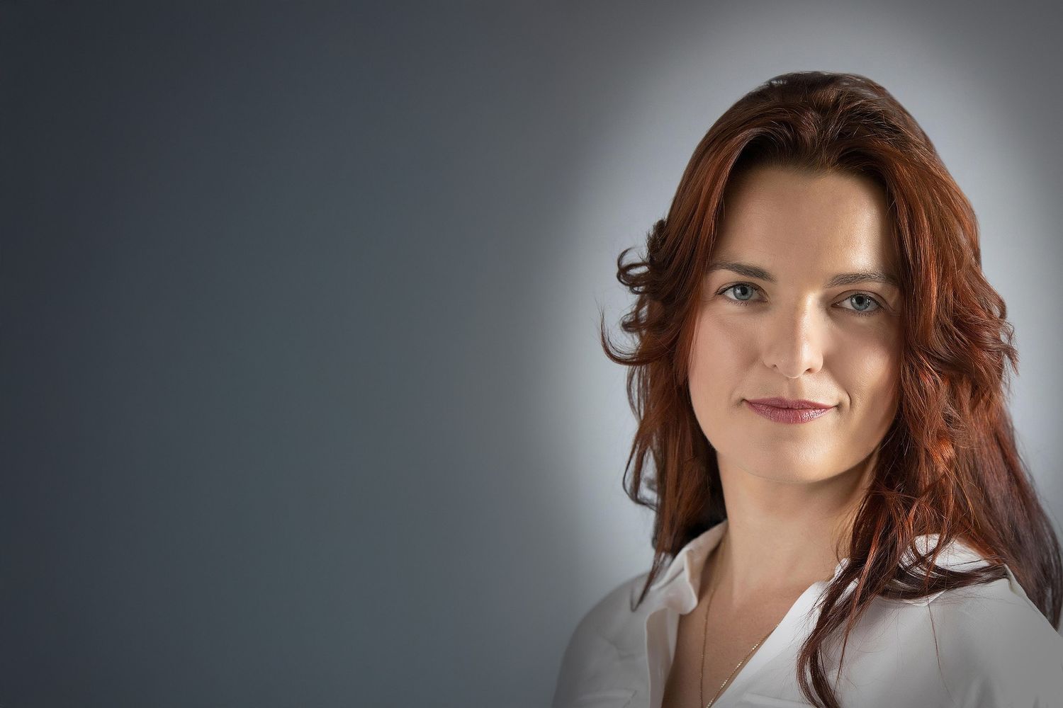 Professional headshot portrait of someone with long red hair wearing a white shirt against a dark gray background.
