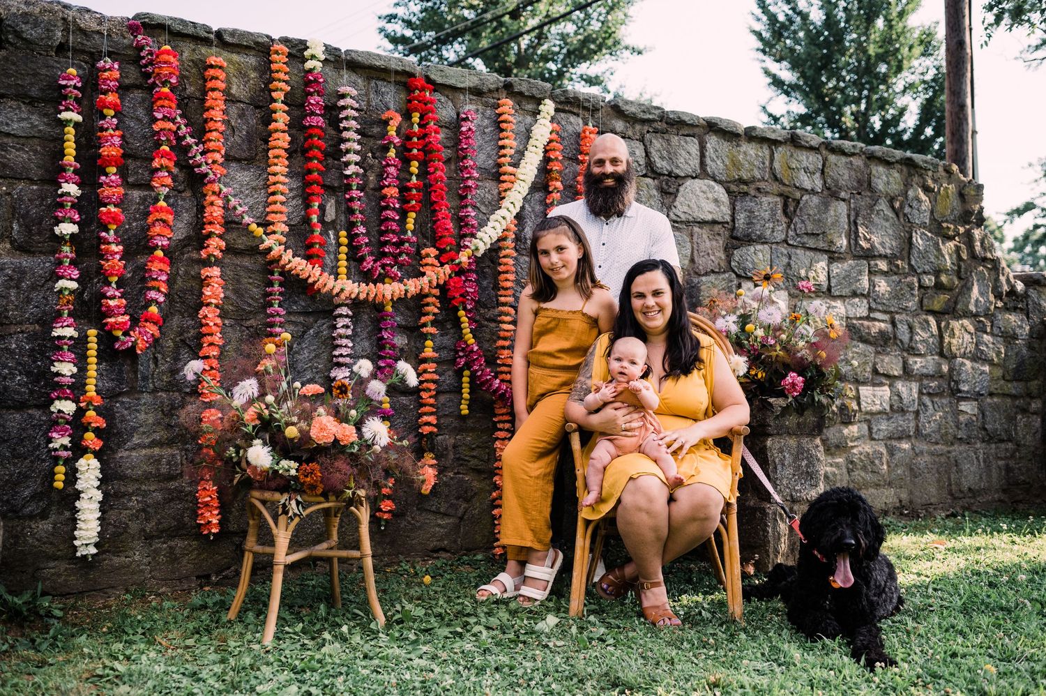 A couple sits on wooden chairs against a stone wall decorated with vibrant orange marigold garlands and a black dog nearby.