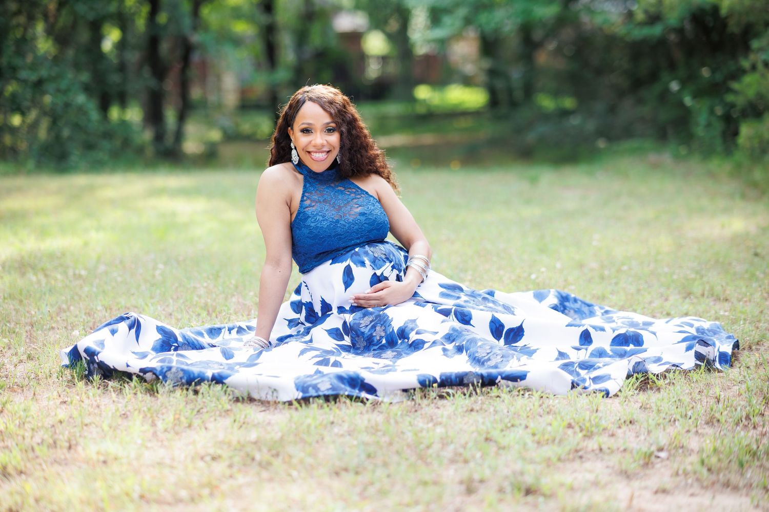A pregnant woman in a flowing blue and white floral dress sits gracefully on grass in a Dallas park during her maternity photo session