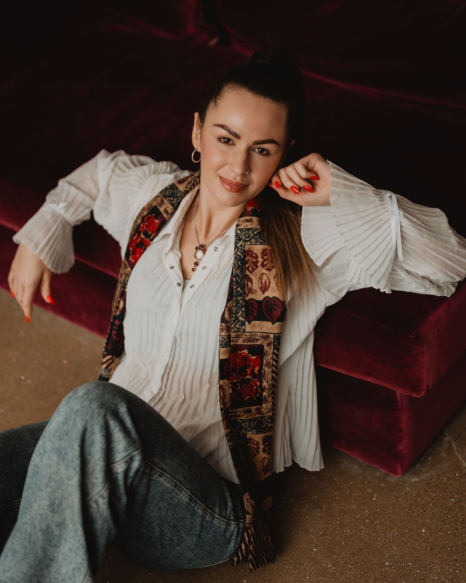A person in a white blouse and patterned vest lounges casually against a red velvet couch in a moody portrait setting.