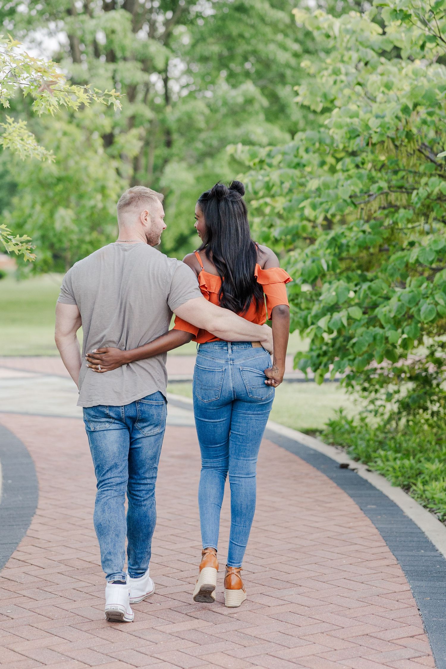 Couple walking together on brick pathway through lush garden setting while holding each other close.