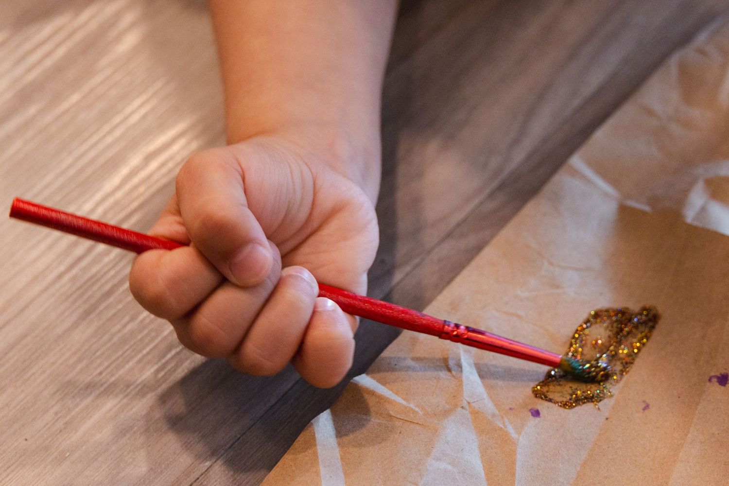Small hand holding a red pencil over a wooden surface while drawing or writing.