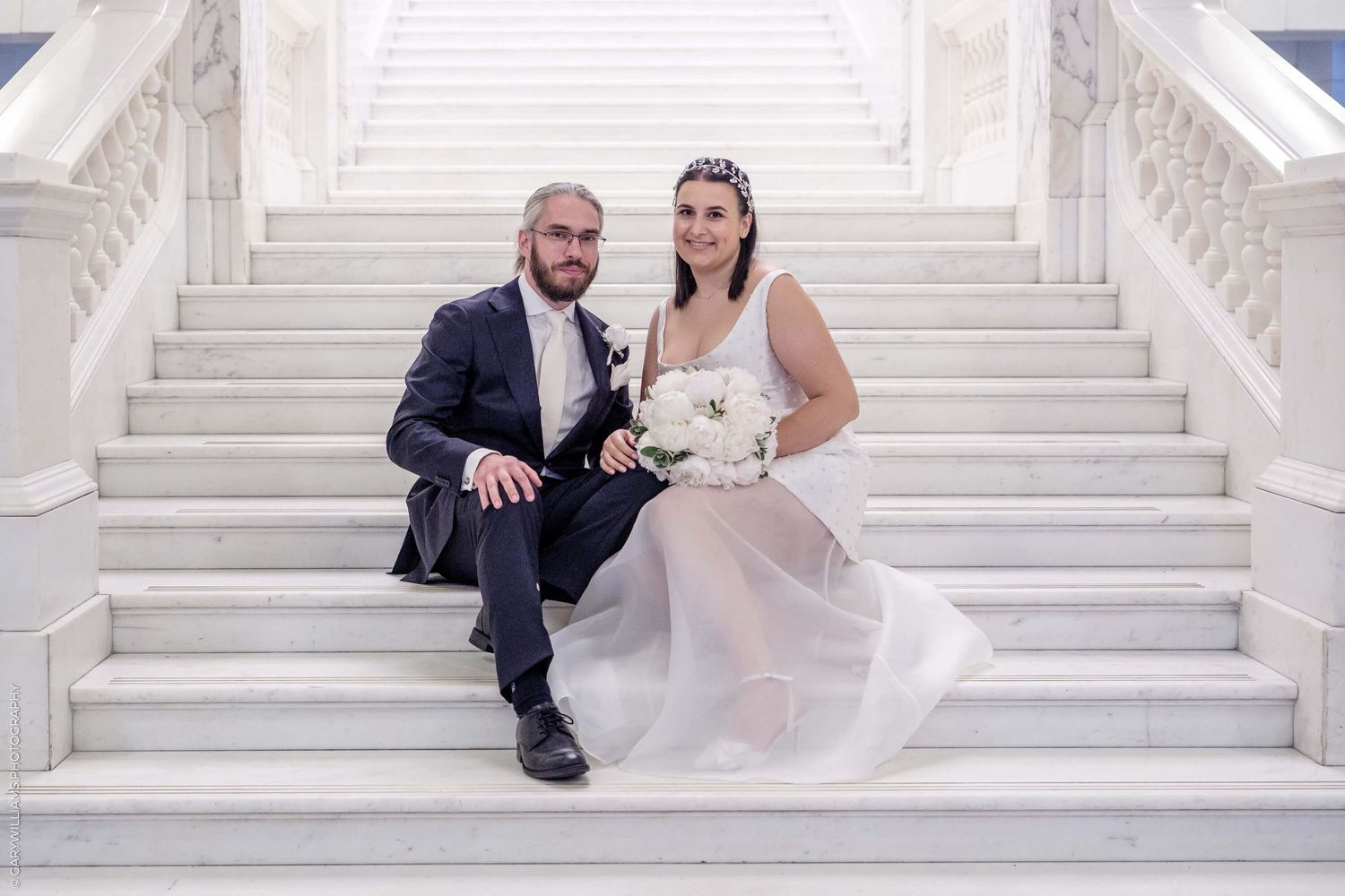 A couple in wedding attire sits together on elegant white marble steps.