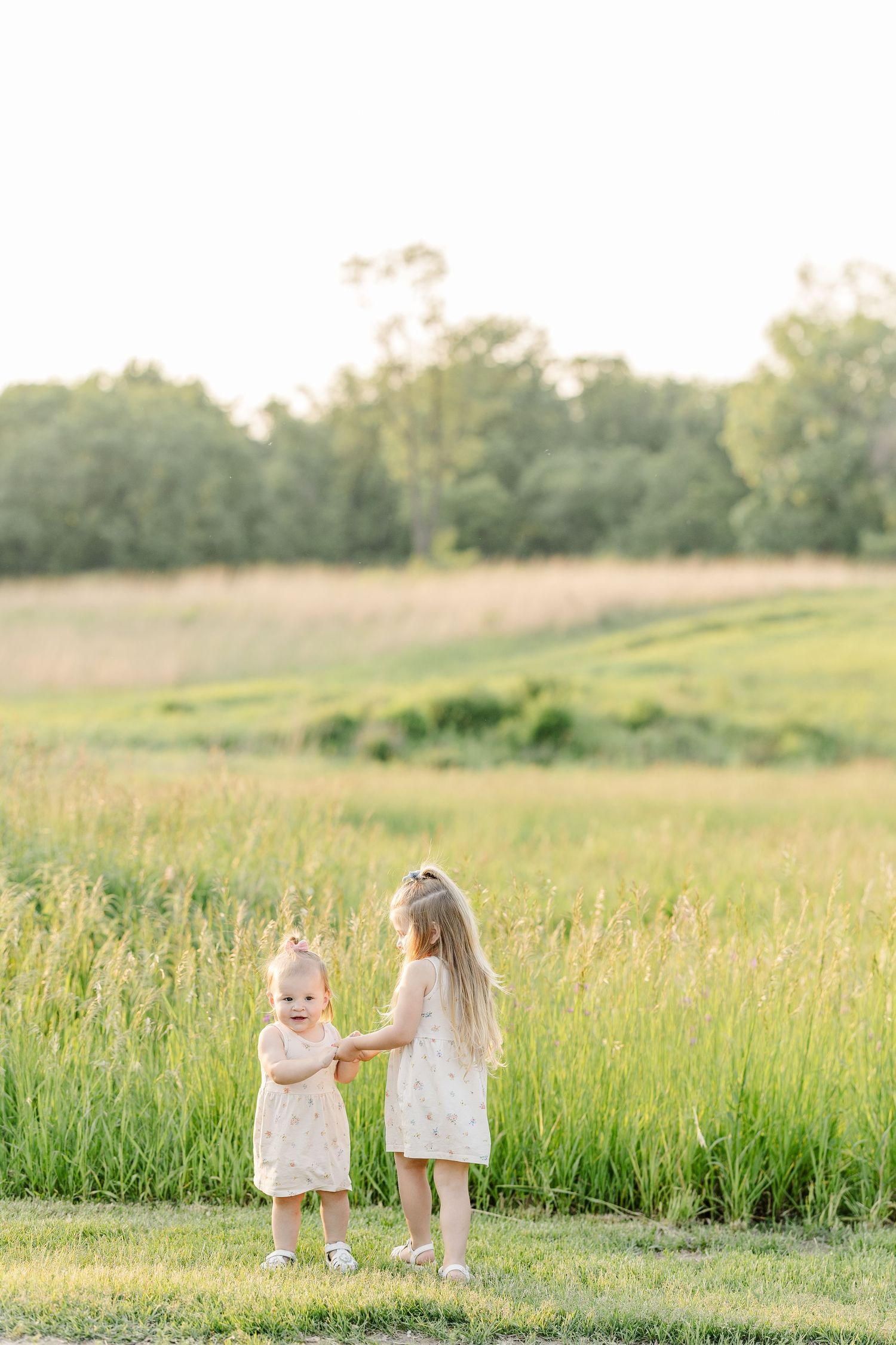 Two children in light colored clothing walking together in a sunlit meadow filled with tall grass at golden hour.