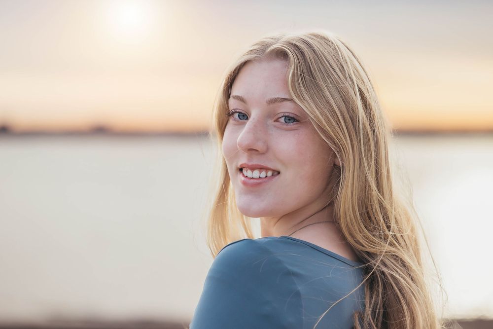 Woman with blonde hair smiles warmly by the water during golden sunset along coastal shoreline.