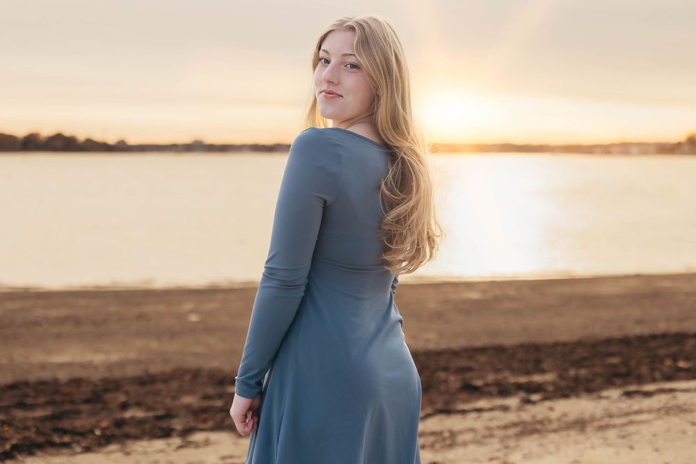 Person in a blue dress stands by a waterfront beach at sunset with warm golden light reflecting on the water.