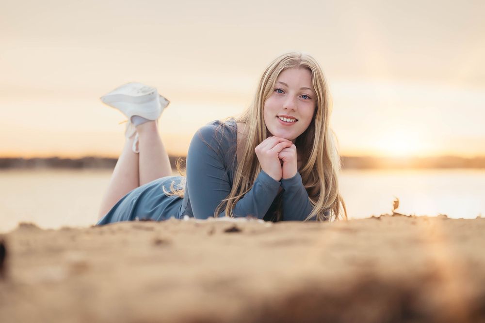 A person sits thoughtfully on a beach during a warm golden sunset with calm ocean waves in the background.