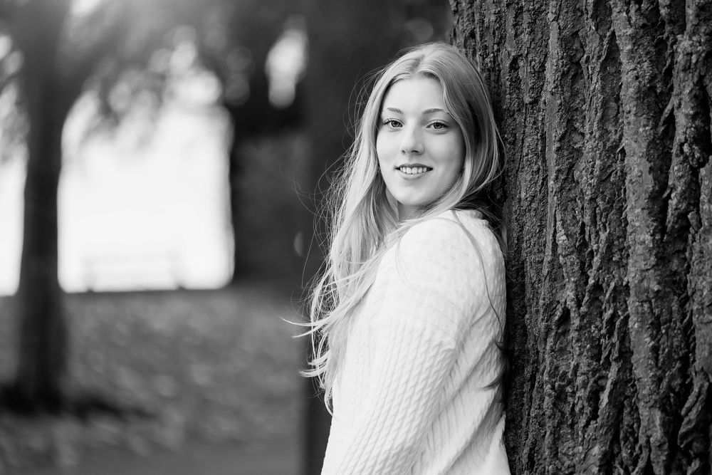 Black and white portrait of person in white sweater leaning against tree trunk with soft natural lighting.
